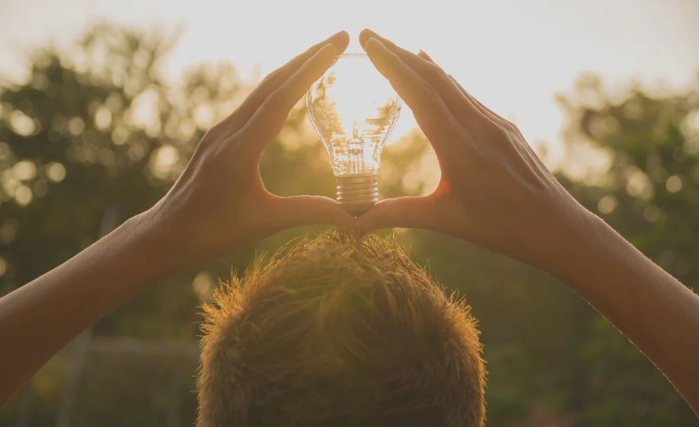 Personne qui tient une ampoule entre ses mains par laquelle passe la lumière pour représenter l'optimisation de sa consommation d'énergie aux moments les plus ensoleillés